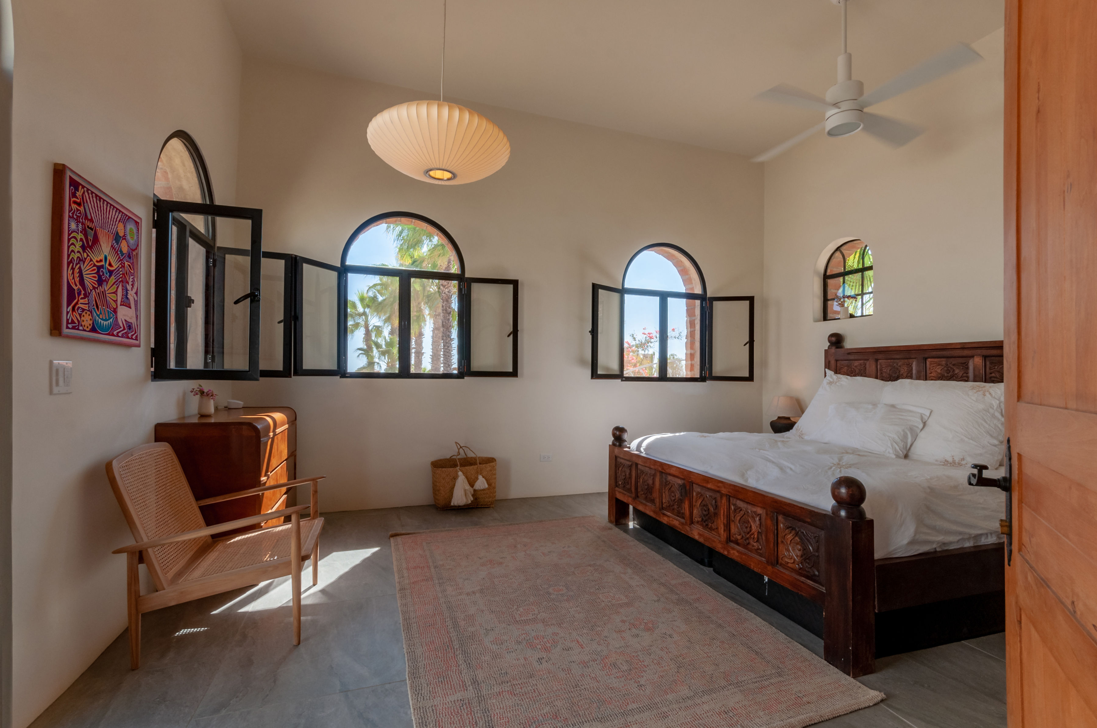 Primary bedroom in the Adobe house with a carved wooden bed, cane chair and arched windows opening onto the palms