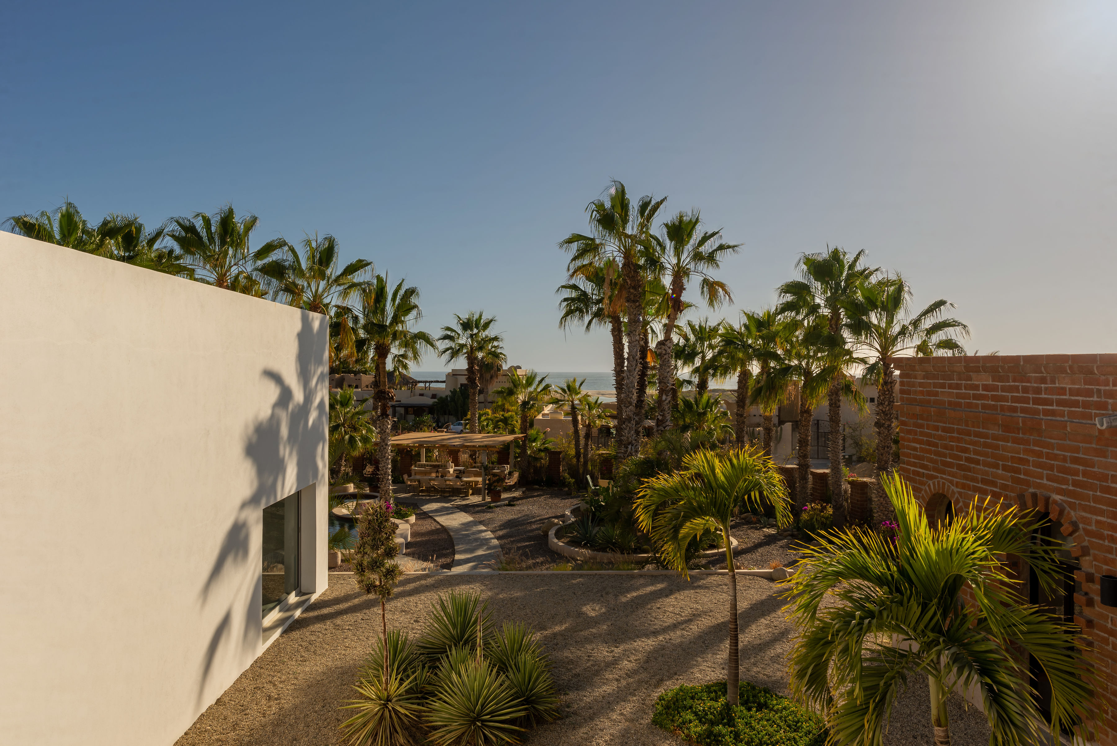 Gravel garden and palms between the white modernist volume and the brick wall, with the ocean beyond