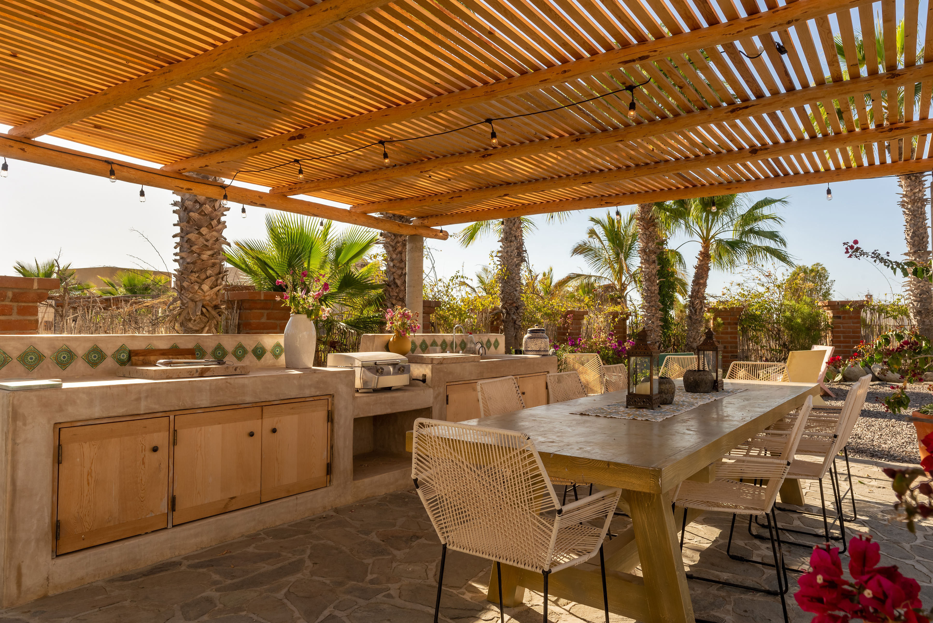 Outdoor kitchen under the slatted pergola: tiled concrete counter with grill and sink, string lights and the long dining table beside it